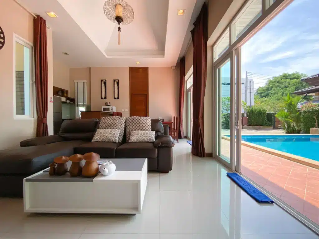 A modern living room with a large sliding glass door opening to a patio with a swimming pool, reminiscent of the upscale Phuket pool villa aesthetic. The room has a dark brown sofa with patterned cushions, a white coffee table with decorative items, and offers views of the blue sky and lush greenery outside. Neutral tones and bright lighting enhance its elegance.