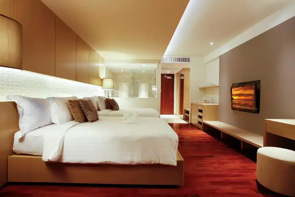 A modern hotel room in Phuket with warm lighting, featuring two neatly made beds with white linens and decorative pillows. A wall-mounted TV is opposite the beds. The room has wooden floors, a small desk area, and a glass-walled bathroom in the background. Minimalist decor and contemporary furnishings.