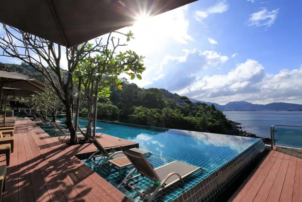 A serene infinity pool at a Phuket pool villa with two sun lounges overlooks a lush, green hillside and a calm, expansive body of water. The pool area is shaded by beach umbrellas and leafy trees. The sun shines brightly in a clear blue sky, creating sparkling reflections on the water's surface.