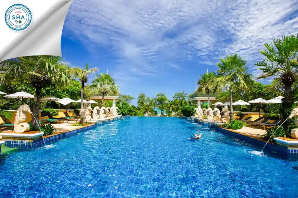 A luxurious, inviting outdoor swimming pool with bright blue water is surrounded by lush green palms at one of the finest hotels in Phuket. White umbrellas and lounge chairs line the sides. Ornate water fountains resembling sculptures add elegance. The sky is clear blue with scattered wispy clouds. SHA logo is visible in the top left corner.