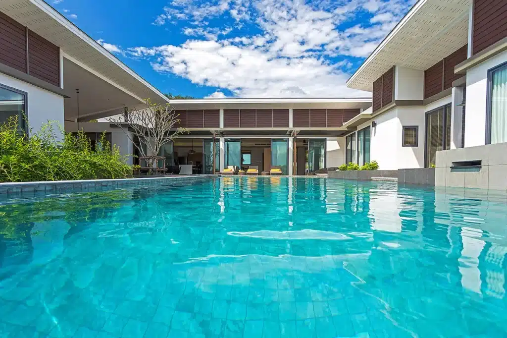 A luxurious modern Phuket pool villa features a serene outdoor swimming pool framed by two white-walled wings of the building. The sky is bright blue with scattered clouds. Lush green plants border the pool area, and poolside seating is visible through the large glass doors of the structure.