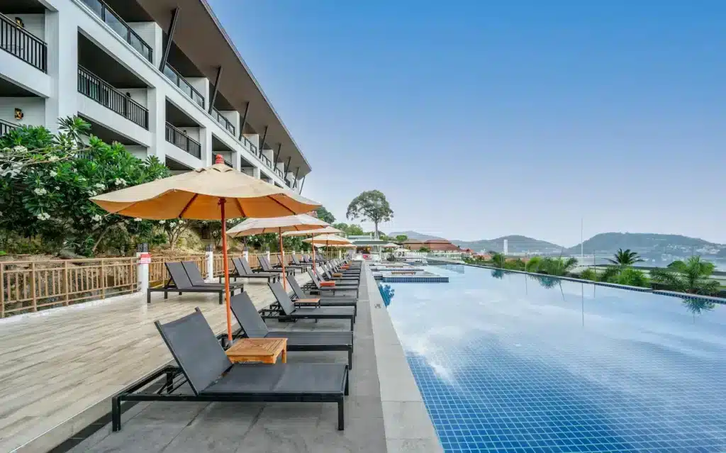 An outdoor infinity pool bordered by black lounge chairs with straw parasols on a tiled deck. The pool overlooks a scenic landscape with rolling hills and buildings in the distance. On the left, there is a large, multi-story building with balconies and surrounding greenery typical of hotels in Phuket. The sky is clear and blue.