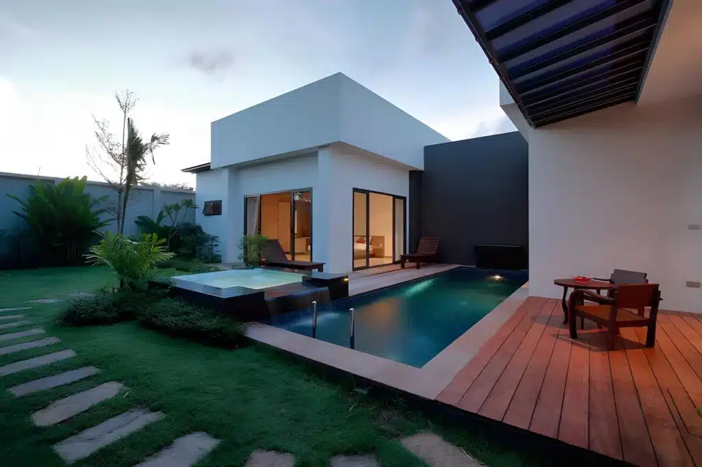 A modern house with a rectangular design features large windows and a minimalist aesthetic. Its outdoor area resembles the elegance of Phuket pool villas, boasting a swimming pool, hot tub, wooden deck with a dining table and chairs, and a lush garden with stepping stones on a well-manicured lawn under evening light.