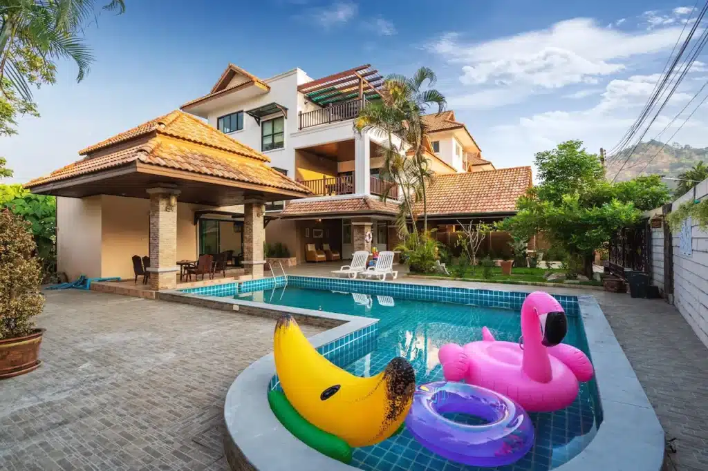 A luxurious backyard of a Phuket pool villa featuring a swimming pool with various inflatable toys, including a banana, flamingo, and donut float. Next to the pool are two white lounge chairs and a patio area with dining furniture. The large, multi-story house has terracotta roofing, balconies, and lush greenery surrounding it.