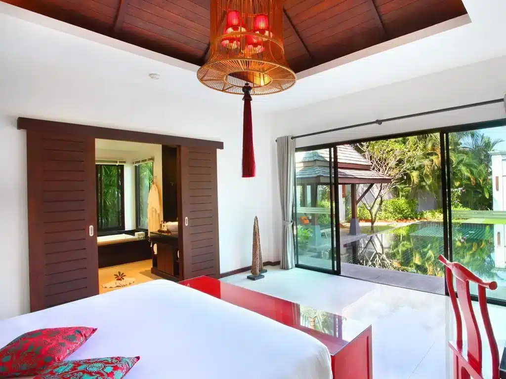 A modern bedroom with an open layout. A large bed with white linens and colorful pillows is in the foreground. Sliding wooden doors reveal a bathroom with a tub. Panoramic glass doors lead to a lush garden with a pool. A red chair sits by the window, and a stylish light fixture hangs from the ceiling.