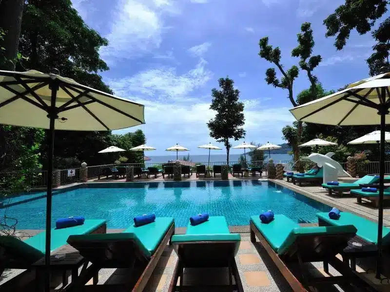 A luxurious outdoor pool area with turquoise lounge chairs and rolled towels, surrounded by large white umbrellas. The pool overlooks a scenic ocean view, framed by trees and clear blue skies. Located among the finest hotels in Phuket, tables and chairs are placed near the poolside stairs with decorative dolphin statues.