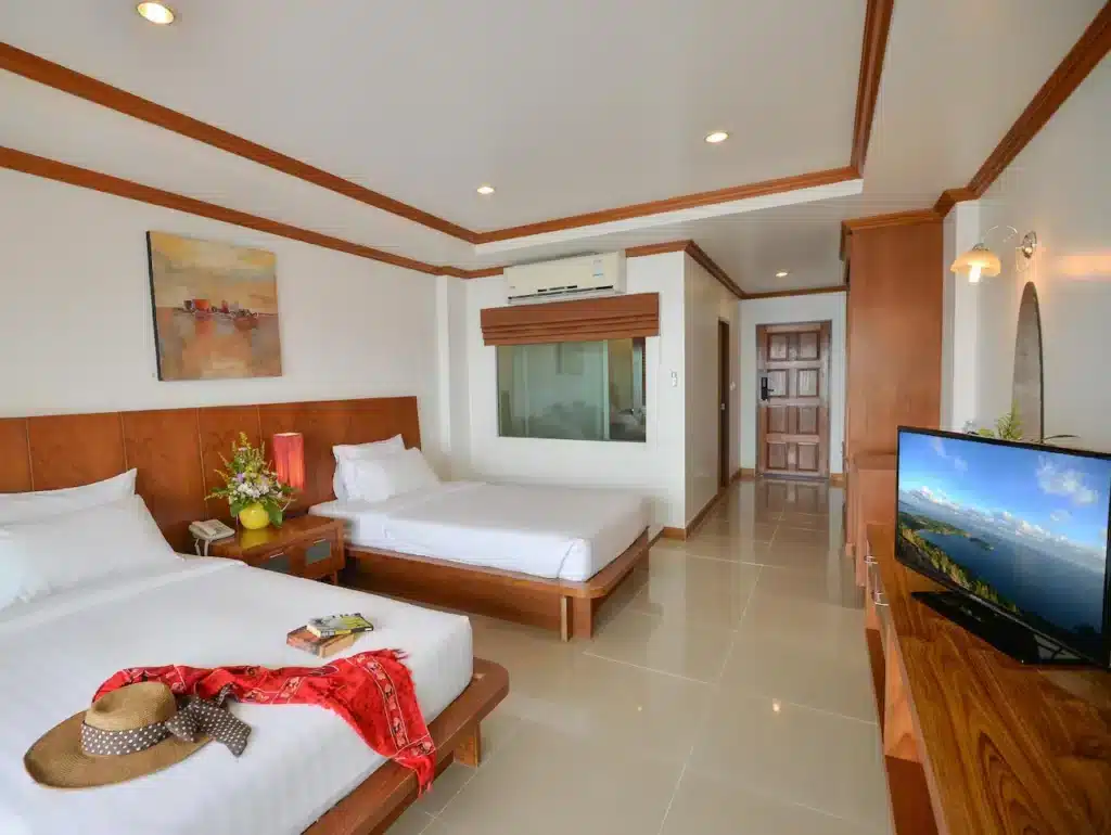 A spacious hotel room with two neatly made beds, wooden headboards, and a red scarf with a hat placed on one bed. The room features a flat-screen TV, a wooden dresser, a large painting on the wall, and a vase of flowers between the beds. With cream-colored walls and tile flooring, it rivals top hotels in Phuket.