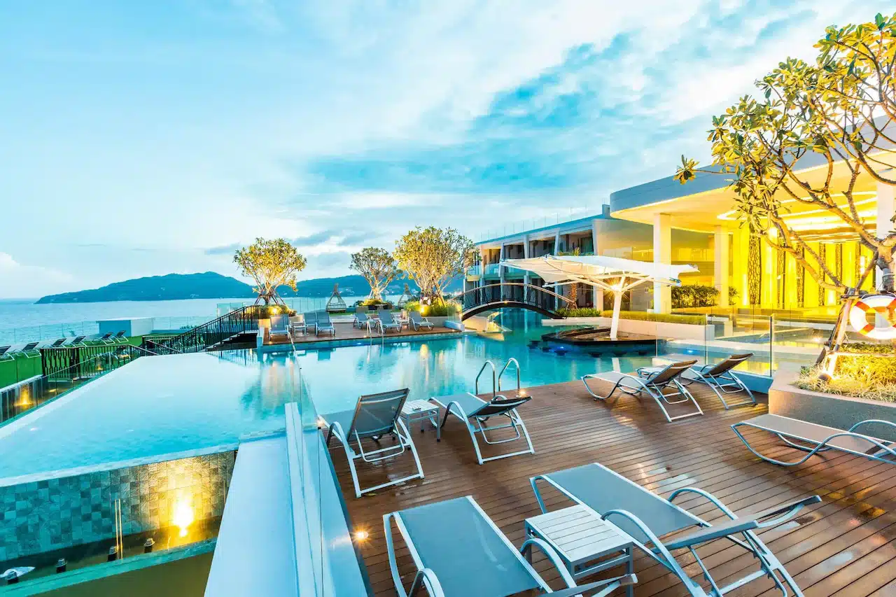 Luxurious beachfront resort featuring an elegant, modern pool area with various lounge chairs and sunbeds on a wooden deck. The pool overlooks a serene ocean with mountains in the background. A small bridge spans the pool, adding sophistication to the relaxed ambiance of our Private Pool Villas in Phuket.