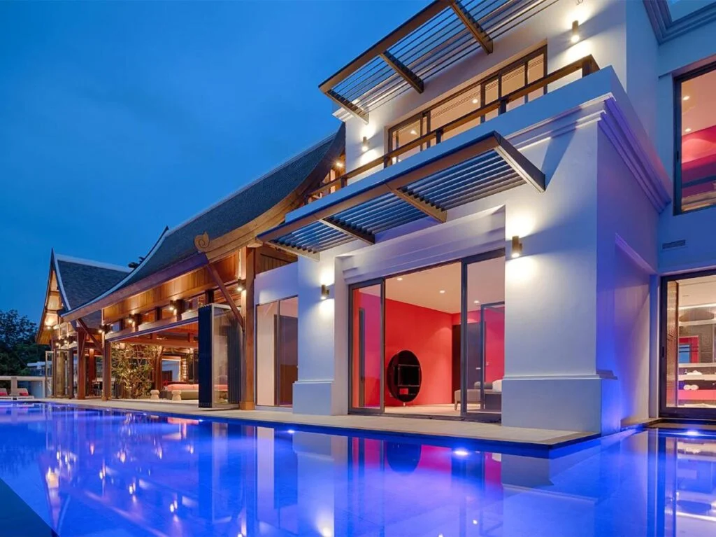 A modern, luxurious house is illuminated during twilight, showcasing a blend of contemporary and traditional architectural elements. A serene pool with glowing blue lights stretches in the foreground, reflecting the house's sleek lines and large windows—reminiscent of exclusive Private Pool Villas in Phuket. The sky is a deep blue, adding to the tranquil ambiance.
