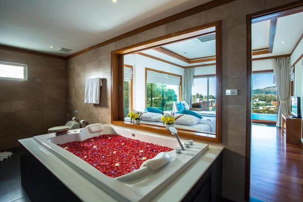 Luxurious bathroom in a Private Pool Villa in Phuket, featuring a large white bathtub filled with rose petals. The room is equipped with modern amenities, including a mounted towel rack. A window offers a view into a plush bedroom with decorative pillows, fresh flowers, and a scenic balcony.