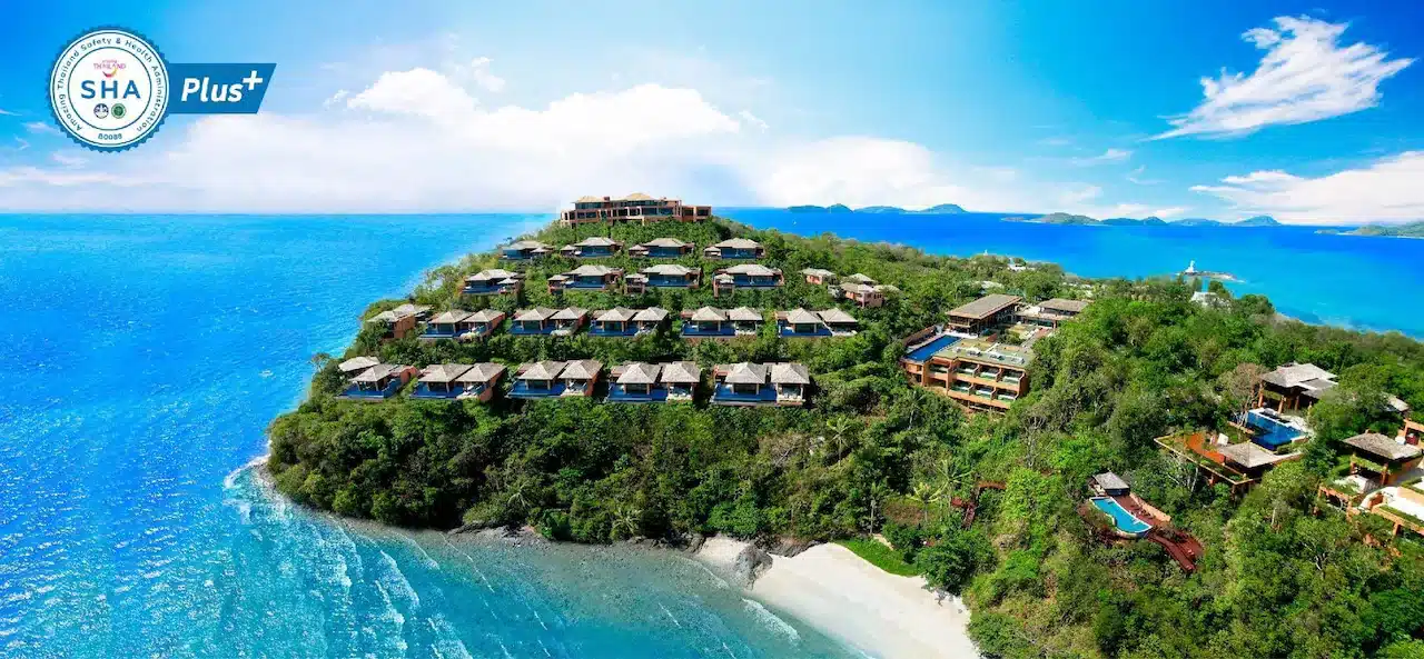 Aerial view of a luxurious seaside resort on a green, hilly peninsula surrounded by bright blue sea. The resort, highlighted in many Phuket travel guides, features an array of cabins and multistory buildings with thatched roofs nestled among lush greenery. The sky is clear with few clouds, and a spacious private beach is visible below.