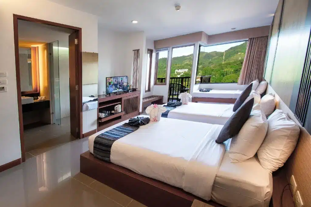 A modern hotel room with two neatly made double beds, each adorned with dark grey accent pillows and folded towels shaped like swans. The room features a flat-screen TV, wooden furniture, large windows with views of lush green hills, an en-suite bathroom with a well-lit vanity area, and offers exclusive access reminiscent of Phuket pool villas.