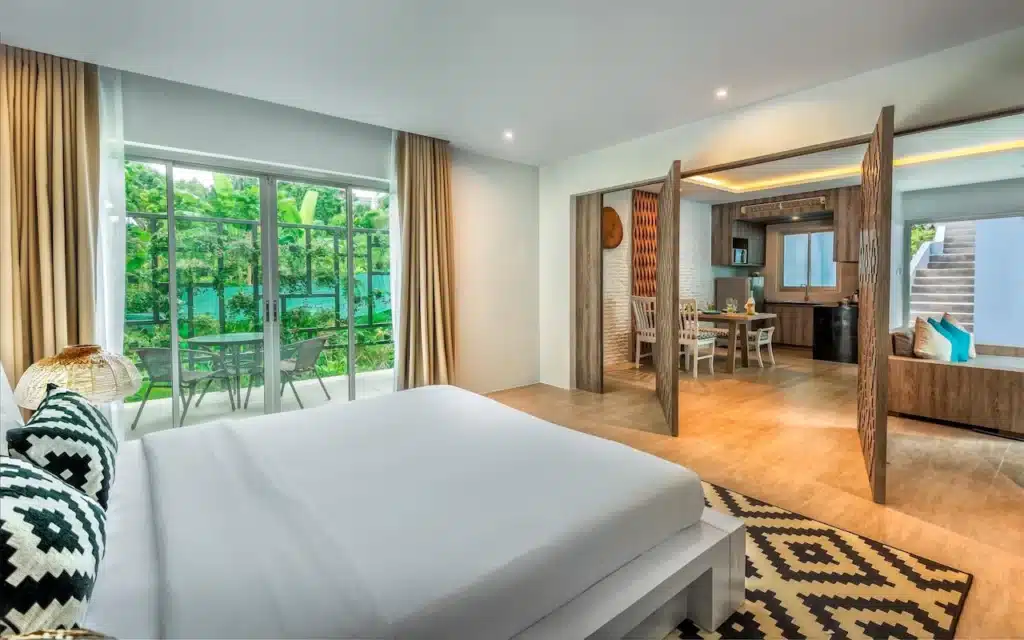 A spacious, modern hotel room featuring a large bed with a white mattress and black-and-white patterned pillows. The room has floor-to-ceiling windows with beige curtains, offering a view of lush greenery. The open-plan area includes a dining table and a kitchen with wooden accents, reminiscent of Private Pool Villas in Phuket.