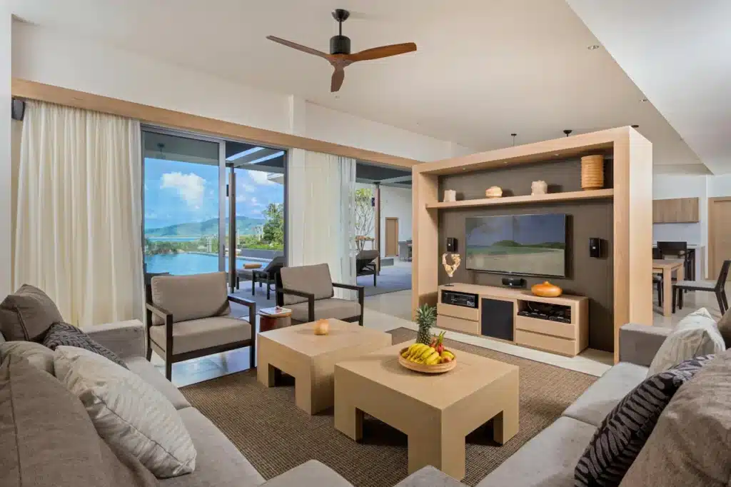 A modern living room in a Phuket pool villa features a large flat-screen TV mounted on a light wood entertainment unit with shelves and decor. Beige sofas with throw pillows face matching coffee tables holding fruit bowls. Large glass doors open to a patio and scenic mountain views.