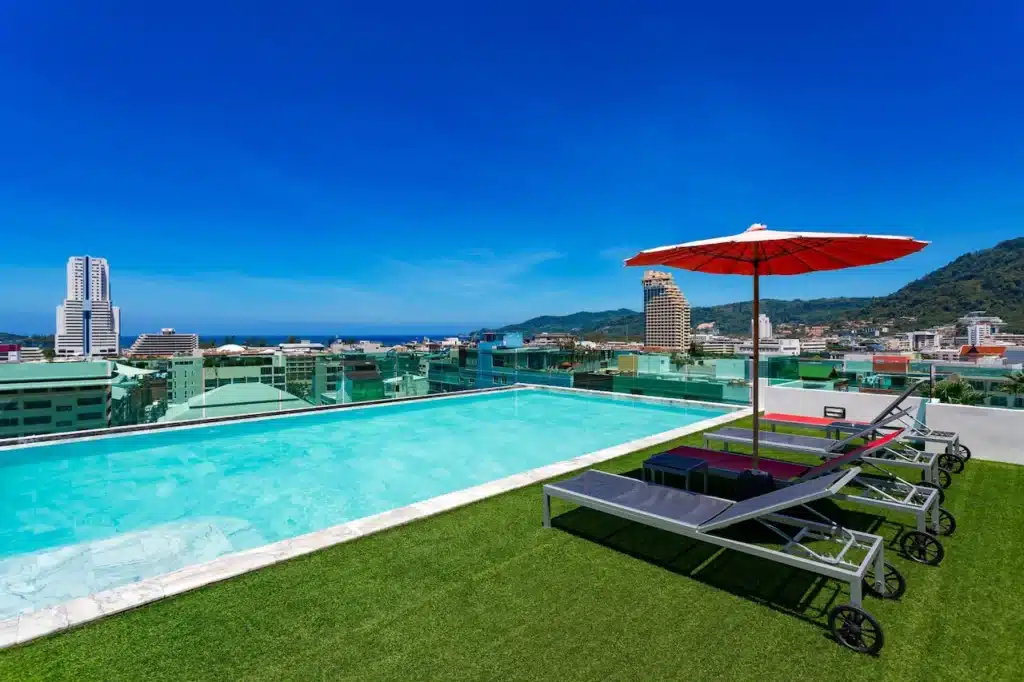 A rooftop swimming pool with crystal clear water and adjacent sun loungers under a red umbrella, sitting on a green artificial lawn. This scene at one of the finest hotels in Phuket overlooks an urban skyline with tall buildings and mountains in the distance under a bright blue sky.