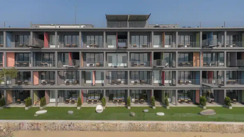 A multi-story modern apartment complex with large glass windows and balconies, situated by the beach, reminiscent of hotels in Phuket. Each unit features outdoor seating areas, and a well-maintained lawn with several lounges and decorative stone spheres graces the front of the building under a clear blue sky.