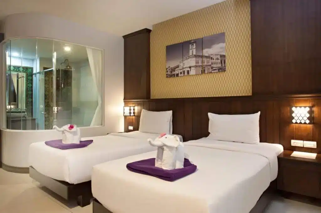 A hotel room with two neatly made single beds adorned with purple towels folded like elephants. Above the headboards, there is a large black-and-white photo of a clock tower. The room, like many hotels in Phuket, features modern decor, warm lighting, and a glass-walled bathroom.