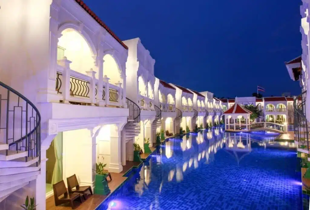 Luxurious resort at night with illuminated arches and a serene blue swimming pool. Curved staircases lead to upper balconies lined with ornate railings. Two wooden chairs are placed on a poolside patio. Red-tiled roofs and palm trees add to the tropical ambiance, embodying the essence of hotels in Phuket.