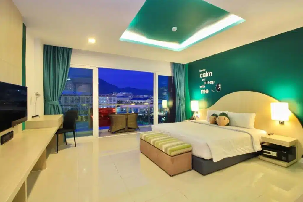 A modern hotel room with white floors and a large double bed adorned with green and beige striped cushions. Green and white walls complement the decor. An illuminated ceiling feature adds ambience. A desk with a flat-screen TV is on the left, and glass doors open to a balcony showcasing Phuket's night view.