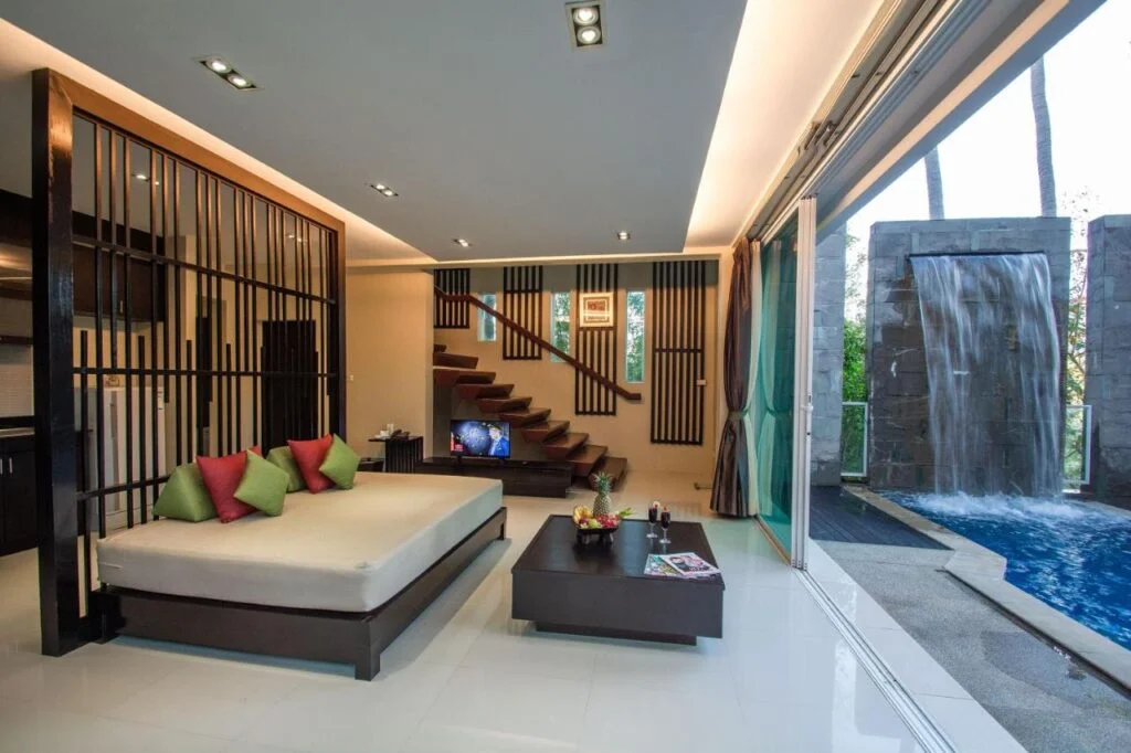 Modern living room with minimalist decor, featuring a white platform bed with colorful pillows, a low coffee table, and an open staircase. Large sliding glass doors reveal the private pool of this Phuket pool villa and a stone waterfall feature outside. Warm ambient lighting enhances the contemporary design.