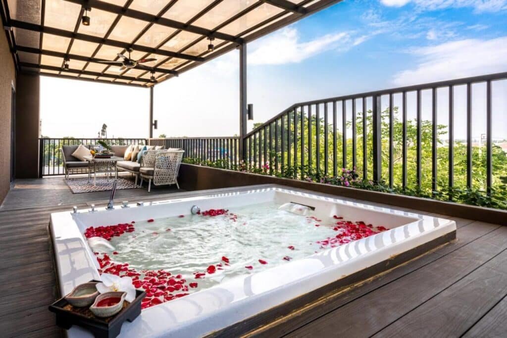 A rooftop deck with a view of the sky and surrounding greenery features a hot tub filled with water and scattered rose petals. There are chairs with cushions, a table with a cloth, and a sofa nearby. Inspired by natural attractions in Bangkok, the area is shaded by a pergola with ceiling fans, providing a relaxed and luxurious outdoor setting.