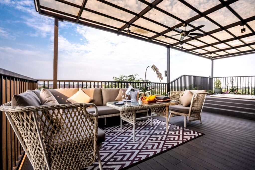 A cozy outdoor patio with wicker furniture arranged around a glass-top coffee table. Beige cushions and pillows accent the seating. The area, reminiscent of the natural attractions in Bangkok, is covered by a clear roof and protected by a black railing. A patterned rug lies beneath the furniture, and a ceiling fan hangs above on this partly cloudy day.