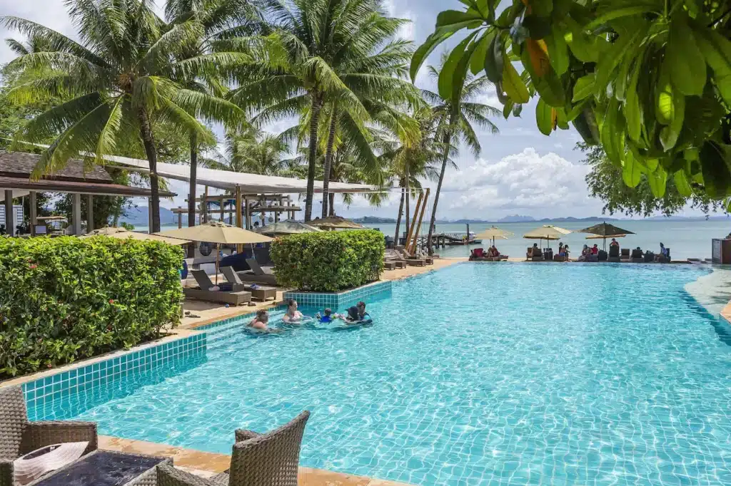 A tropical resort scene features a large, inviting swimming pool surrounded by palm trees and lounge chairs under yellow umbrellas. Several guests enjoy the pool while others relax on loungers. Beyond the pool, a serene ocean view extends to the horizon under a partly cloudy sky, typical of Private Pool Villas in Phuket.