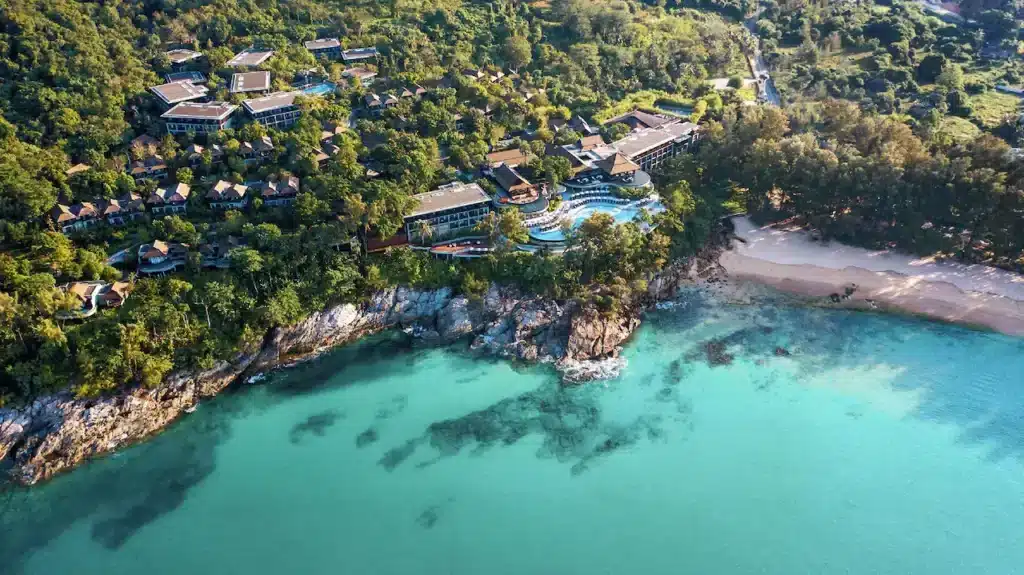 Aerial view of a coastal resort surrounded by lush greenery, located on a rocky promontory with white sandy beaches and clear turquoise waters. Much like the hotels in Phuket, the resort consists of various buildings, pools, and pathways that blend into the natural landscape, with a forested area in the background.