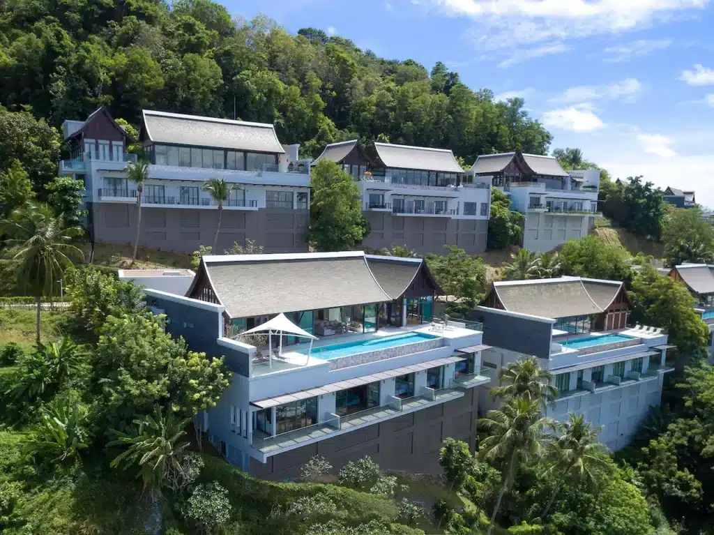 A luxurious hillside resort features modern villas with large windows and Private Pool Villas in Phuket overlooking lush greenery and tropical trees. The buildings have sloped roofs and spacious balconies. The resort offers stunning views of the landscape under a clear blue sky.