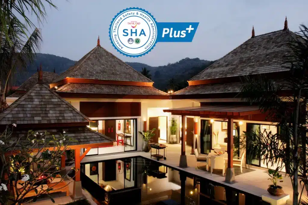 An inviting Phuket pool villa at dusk, featuring traditional Thai architecture with pointed roofs and warm lighting. The villa surrounds a reflective pool, accented by lush greenery. A "SHA Plus+" certification logo is prominently displayed, indicating high health and safety standards.