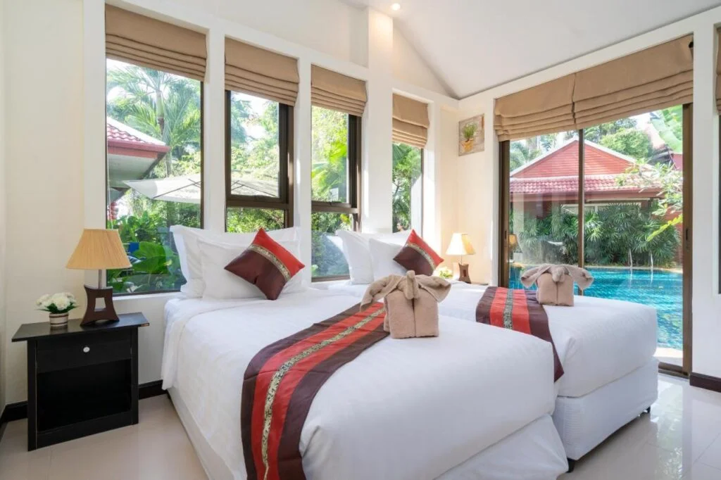 A bright hotel room features two neatly made twin beds with white linens and accent pillows. Towel swans are placed on each bed. The room has large windows with tan blinds, offering a view of a stunning Phuket pool villa and lush greenery outside. Two bedside tables with lamps and a flower vase are beside the beds.