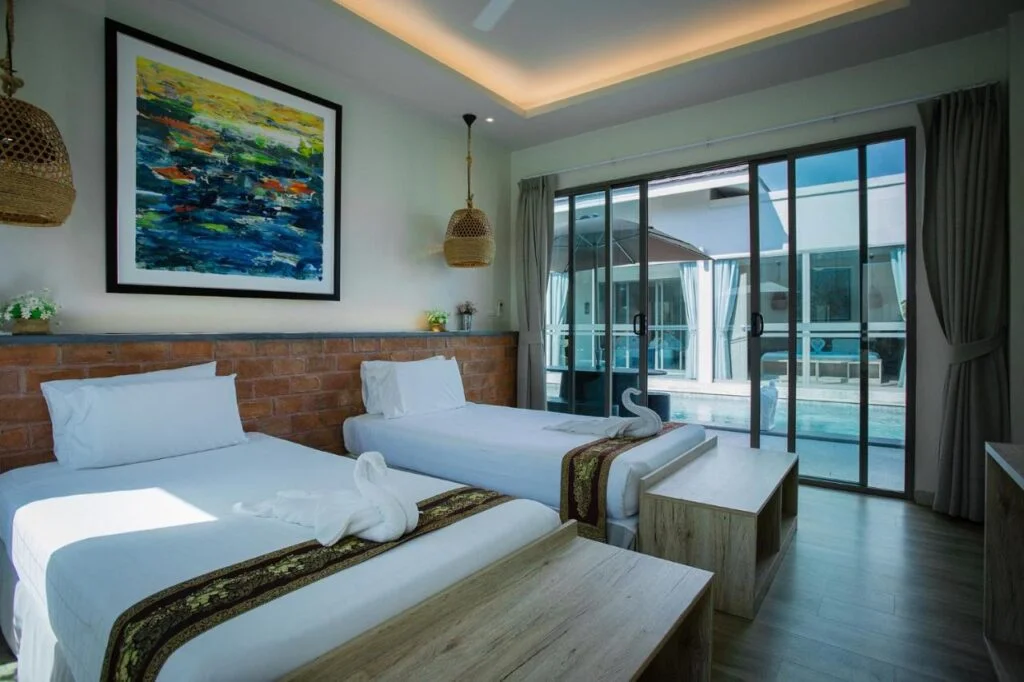 A modern hotel room features two single beds with white linens and decorative pillows. Above the headboard, a colorful abstract painting hangs on a brick wall. The room is decorated with wicker hanging lamps and wooden furniture. Floor-to-ceiling sliding glass doors open to a stunning Phuket pool villa area outside.