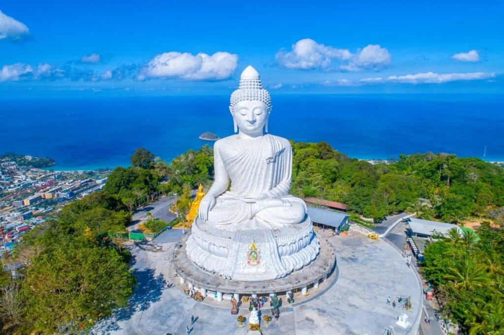 Aerial view of the Big Buddha statue in Phuket, Thailand. The massive white marble statue sits atop Nakkerd Hill, surrounded by lush greenery with a view of the ocean and scattered clouds in the blue sky. A few people are seen at the base of the statue, with nearby hotels in Phuket offering stunning vistas.