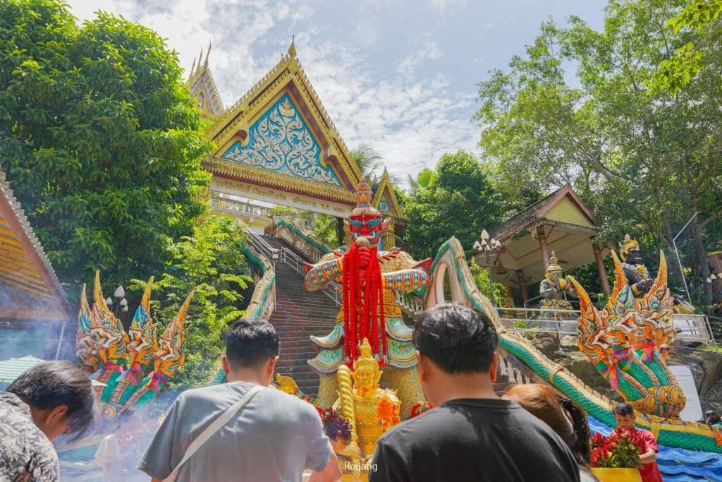 People stand in front of a colorful temple adorned with intricate statues and vibrant decorations. A prominent, ornate dragon statue with red, green, and gold details takes center stage. Surrounded by lush greenery, this traditional temple is a serene escape for visitors staying at hotels in Phuket.