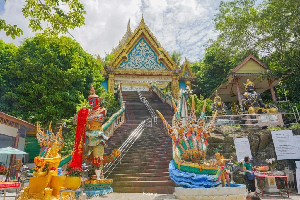 A colorful temple entrance featuring ornate statues and intricate carvings welcomes visitors. A large staircase leads up to a decorated gate, flanked by statues of warriors and mythical creatures with multiple heads and arms. Lush greenery surrounds the area, with signs providing information about nearby hotels in Phuket.