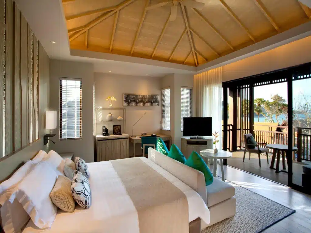 A well-lit bedroom with a high, curved wooden ceiling. It features a large bed with white linens and multiple pillows. The room includes a flat-screen TV, a small dining table with chairs, and a minibar. Sliding glass doors open to a spacious balcony of your Private Pool Villa in Phuket, offering views of the ocean and palm trees.