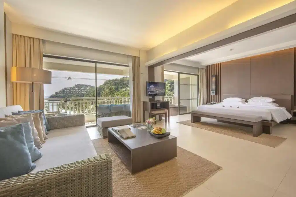 A modern hotel room features a king-size bed, a living area with a wicker sofa, a coffee table, and a TV. Large windows and a sliding glass door offer a scenic view of lush hills and a cable car. Decorated in neutral tones with light wood accents, this example of top Hotels in Phuket includes a fruit basket on the coffee table.