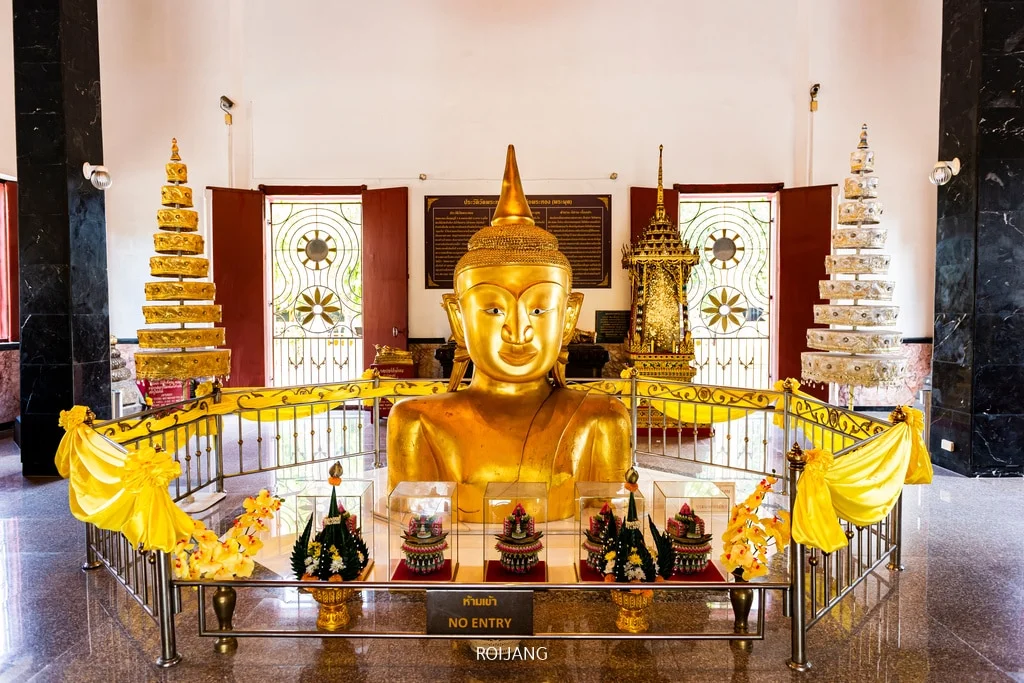 A golden Buddha head statue is prominently displayed in a serene, indoor Temples in Phuket setting reminiscent of the spiritual tranquility often found in high-end hotels in Phuket. The statue is enclosed by a metal barrier adorned with yellow fabric and vibrant floral arrangements, with intricate gold structures and two elaborately decorated doors visible in the background.