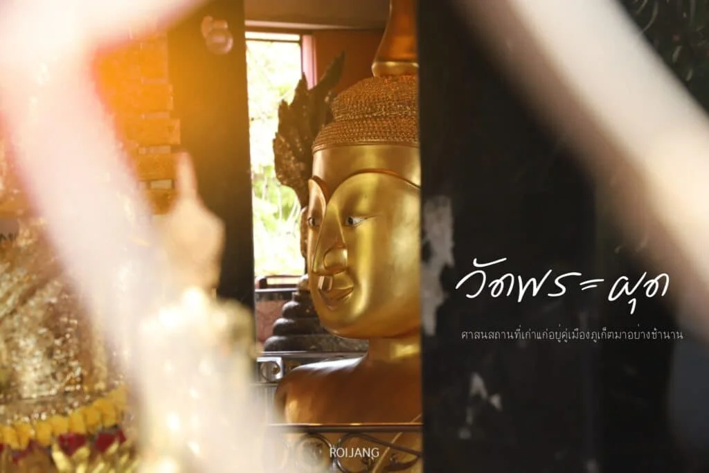 Close-up of a golden Buddha statue in a Thai Temples in Phuket, partially obscured by a white blurry object in the foreground. Thai text is visible on the right side of the image, set against a dark background. The scene is softly lit with warm, ambient light, reminiscent of serene evenings in hotels in Phuket.