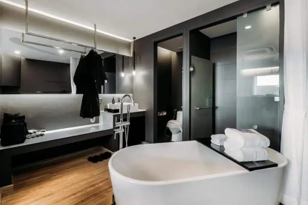 A modern bathroom reminiscent of hotels in Phuket, featuring a freestanding white bathtub with stacked towels and a black tray. Behind the tub, there's a vanity with a mirror and a hanging black robe. The room also includes a glass-enclosed shower, separate toilet area, wood flooring, and dark-toned walls.