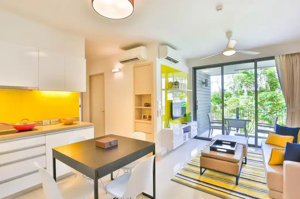 A modern apartment with an open-plan layout features a kitchen, dining, and living area. The kitchen has white cabinets and a yellow backsplash. The dining area has a black table and white chairs. The living area includes a beige sofa, striped rug, and large windows leading to a balcony with garden views reminiscent of hotels in Phuket.