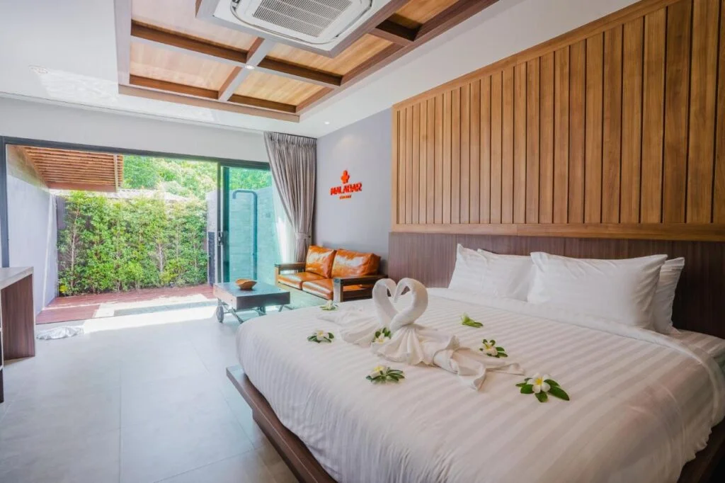 A Private Pool Villa in Phuket features a large bed adorned with white linens, two towel swans, and flower petals. The room has wooden panel accents, a glass coffee table, and an orange sofa. Glass doors open to an outdoor area with green foliage and wooden decking. The ceiling has wooden inlays and an air conditioner.