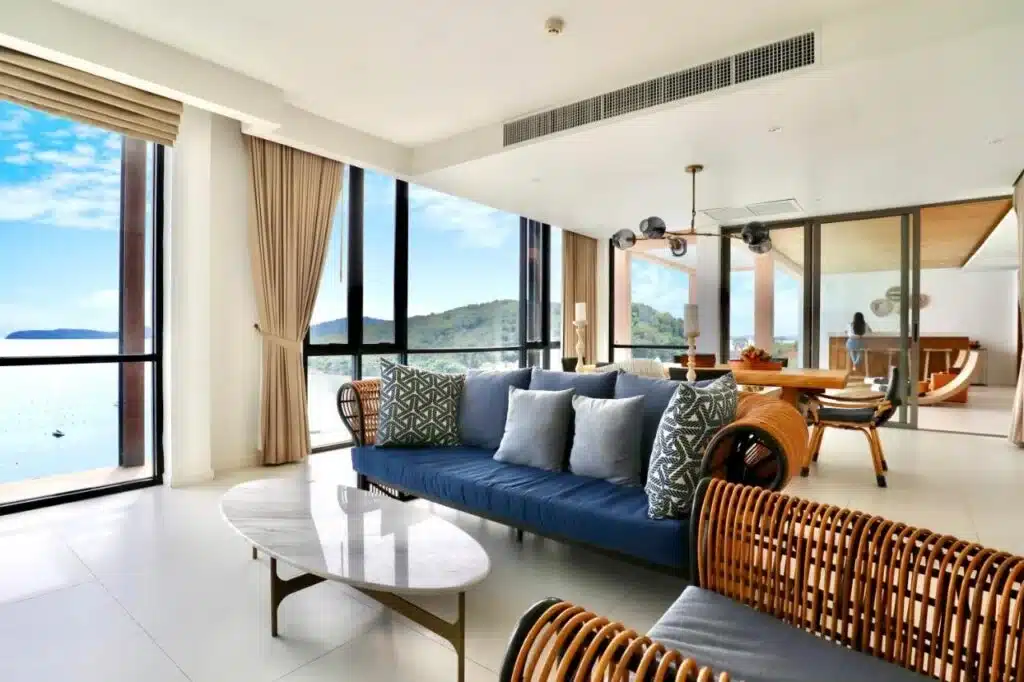 A modern, airy living room with large floor-to-ceiling windows offering a view of a lake and green hills reminiscent of the serene landscapes seen at hotels in Phuket. The space features a navy blue sofa with patterned and plain cushions, wooden rattan chairs, and a marble coffee table. Curtains are half-drawn, and a dining area is visible in the background.