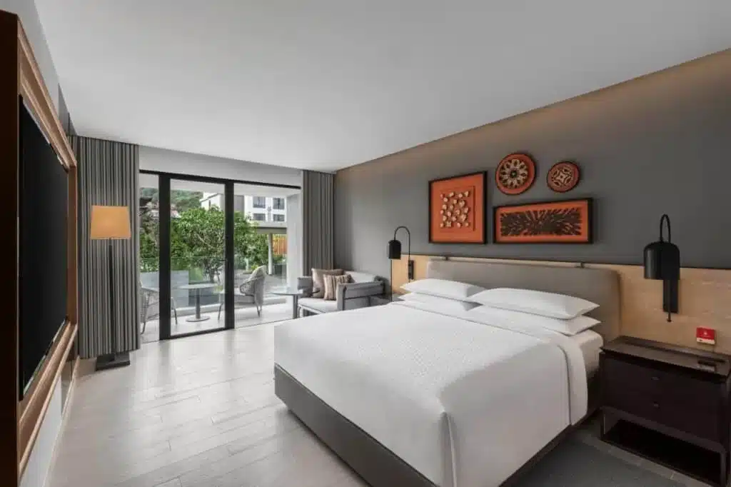 Modern hotel room with a large bed, dark grey accent wall, and wooden floor. Above the bed are three decorative artworks. The room features a floor-to-ceiling window with grey curtains, leading to a balcony with an outdoor seating area. Popular among hotels in Phuket, it includes a dark bedside table, lamp, and wall-mounted TV.