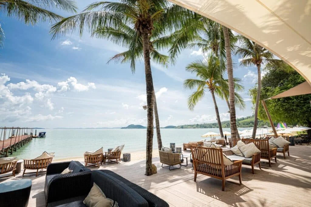 A beachside lounge area with comfortable wooden seating and cushions under tall palm trees. Nearby, a wooden footbridge extends into the calm, turquoise sea. The sky is clear with a few clouds, and the scenic backdrop includes distant hilly islands. Private Pool Villas in Phuket offer similar serene experiences with added luxury.