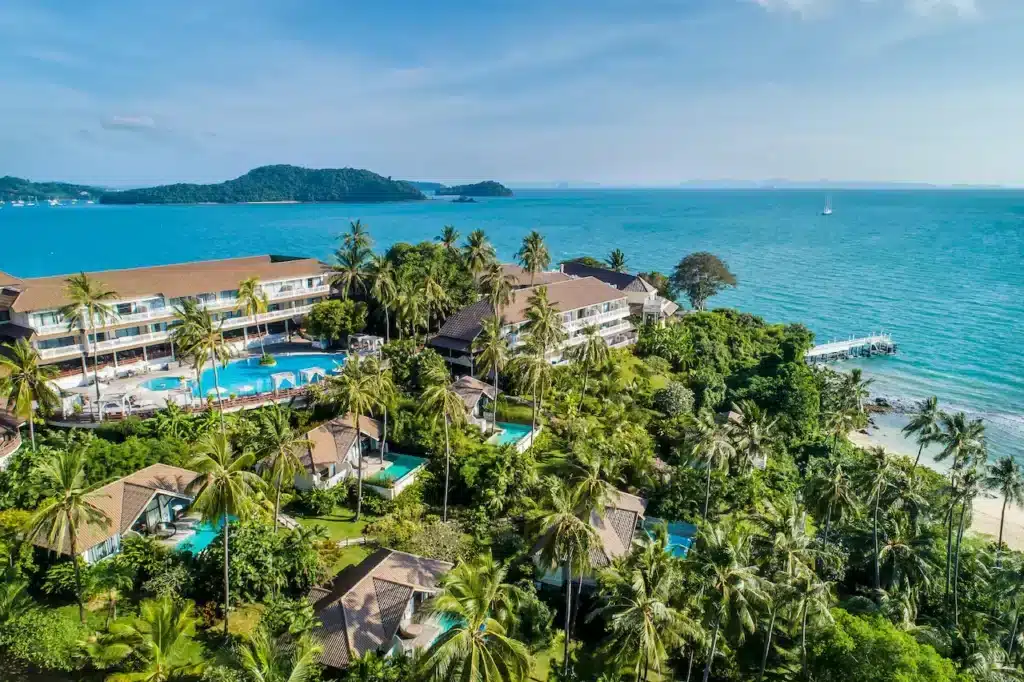 Aerial view of a tropical resort in Phuket featuring multiple buildings surrounded by lush greenery and palm trees. The resort includes outdoor pools and cabanas, with a stunning ocean backdrop. A long pier extends into the clear blue water, and small islands are visible in the distance.