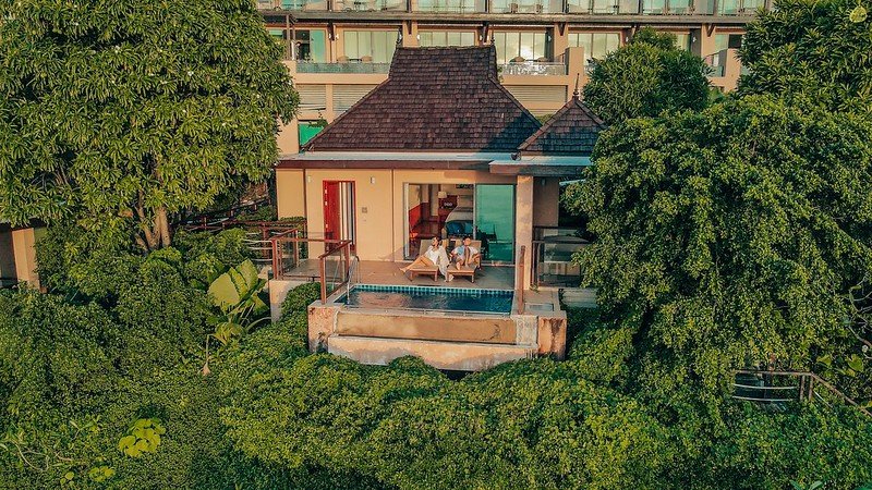 A cozy villa with a small private pool is nestled amidst lush greenery. This Phuket pool villa has a shaded patio with a triangular roof, overlooking dense foliage. Large glass doors offer a view of the interior, and a multi-story building is visible in the background. Two people sit at a table on the patio.