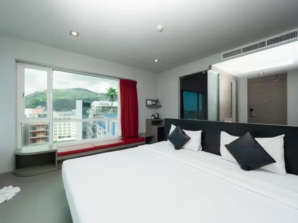 A modern hotel room in Phuket with a large bed topped with white linens and black accent pillows is shown. The room features a window with a scenic view of green hills and buildings, red drapes, a built-in window seat, minimalist furnishings, and a sleek bathroom entrance.