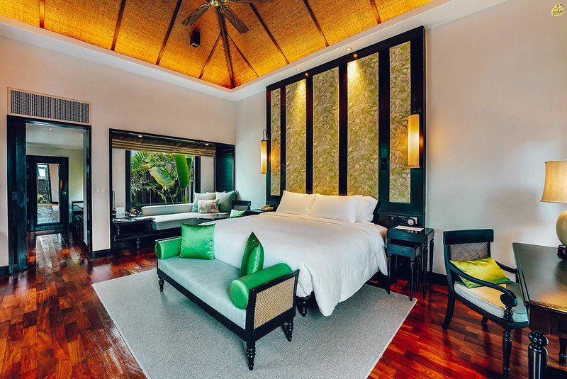 A spacious, luxurious bedroom with hardwood floors in one of the Private Pool Villas in Phuket. The room features a large bed with white linens, flanked by two hanging lights and a decorative wall behind. At the foot of the bed, there is a green-cushioned bench and a chaise lounge next to a window with views of tropical plants.