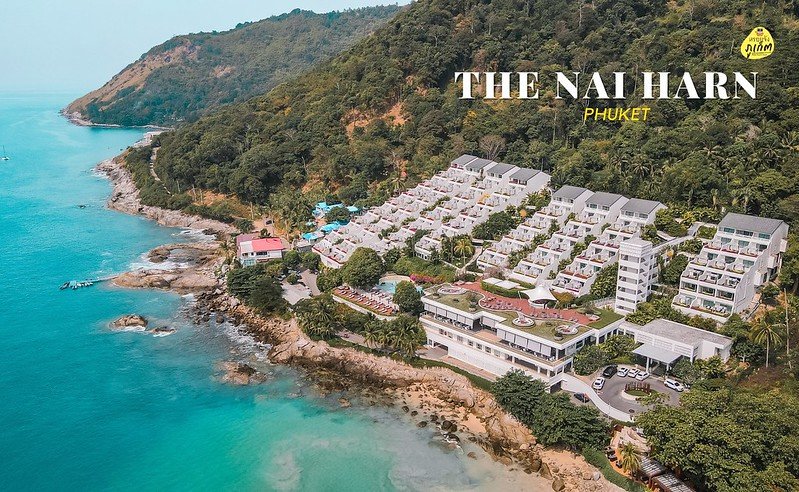 Aerial view of The Nai Harn resort in Phuket, nestled on a verdant hillside overlooking the turquoise sea. The image showcases multiple white buildings with balconies, a pool area, and lush greenery surrounded by a rugged coastline. The logo and text "The Nai Harn Phuket" are superimposed. Ideal for those seeking luxurious hotels in Phuket.